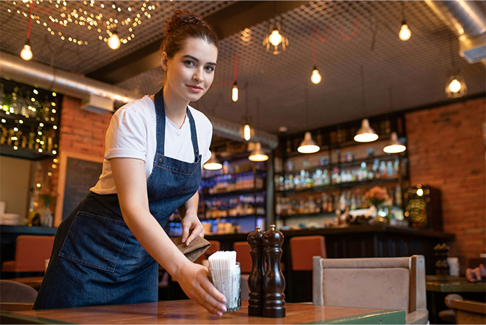 Young waitress in a cafe setting up a table, wearing a white shirt and apron, under warm lighting.
