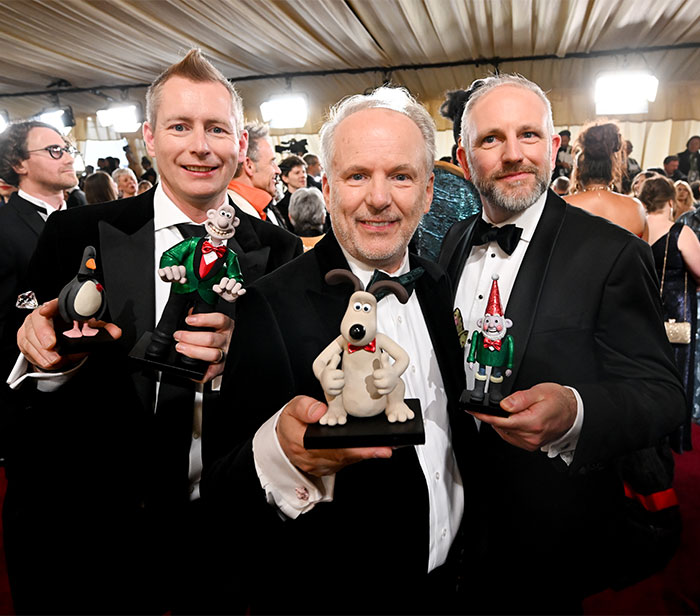 Three men in tuxedos holding animated character models backstage at the Oscars 2025.