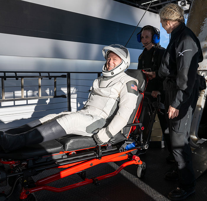 NASA astronaut returns to Earth, smiling in space suit, attended by crew on a stretcher.
