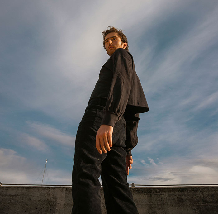 Young man in black clothes stands against a cloudy sky, embodying cultural differences in style and expression.