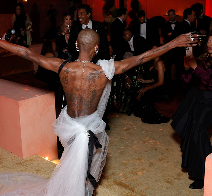 Backstage moment at Oscars 2025: person with tattoos in a unique outfit, engaging with the audience.