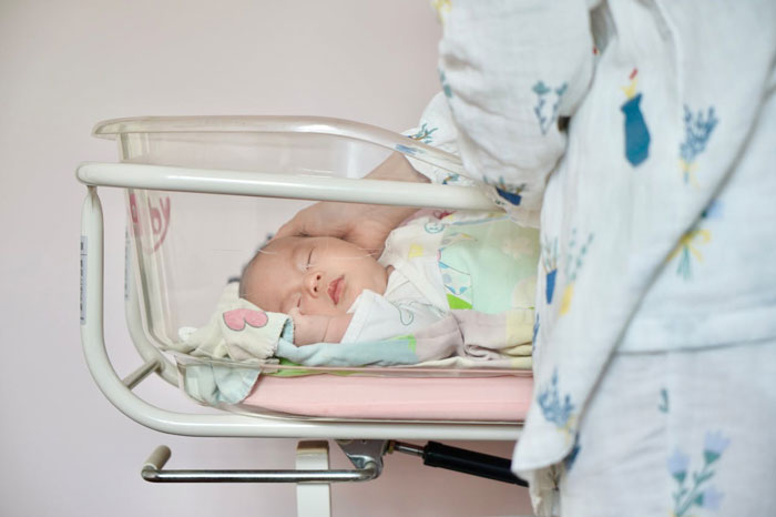 Newborn sleeping in a hospital crib, gently being touched by a person in pajamas.