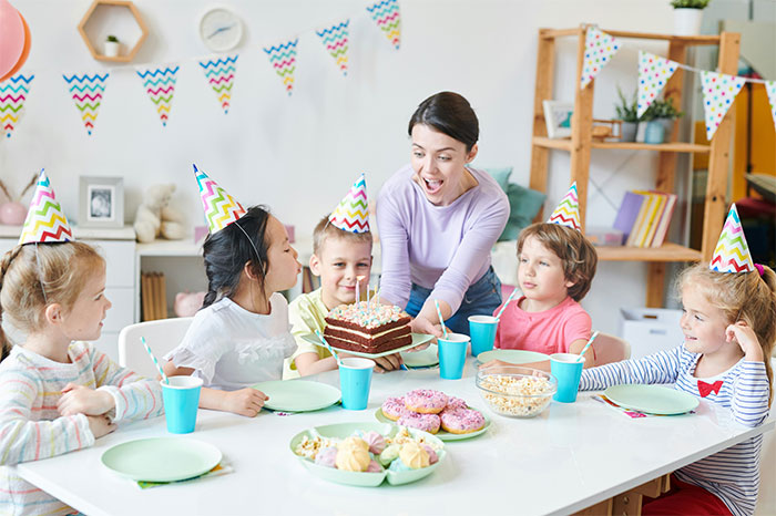 Children celebrate a birthday party with colorful hats, cake, and snacks, while a mom brings a chocolate cake to the table.