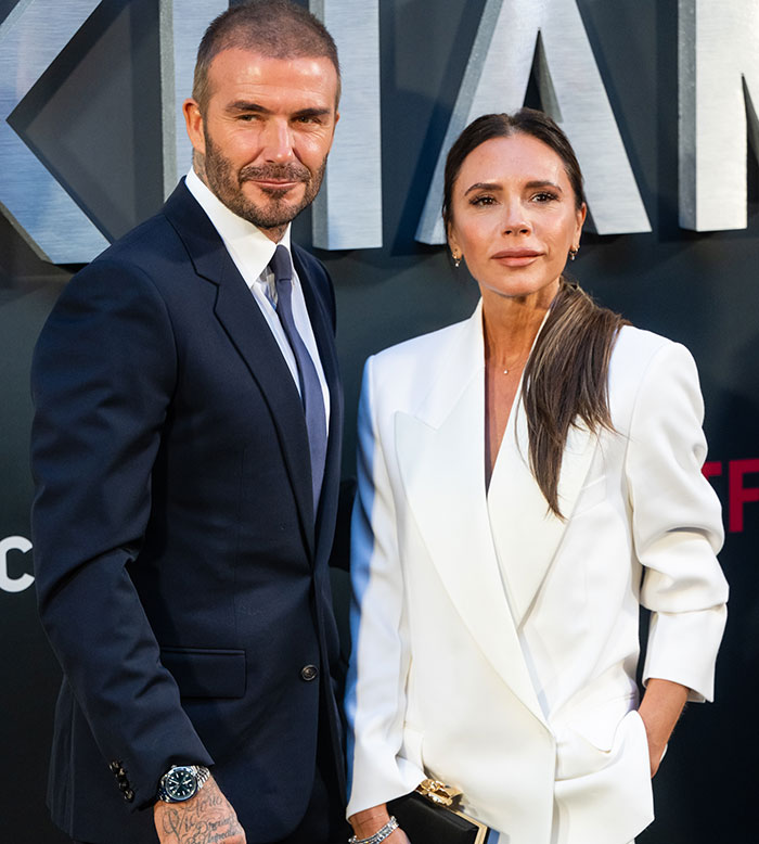 David Beckham at an event in a suit, standing with Victoria Beckham in a white blazer.