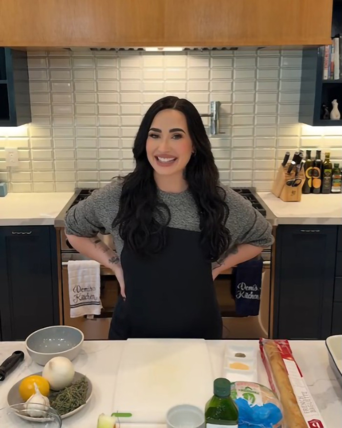 Demi Lovato smiling in a kitchen, wearing a black apron, showcasing weight loss transformation.