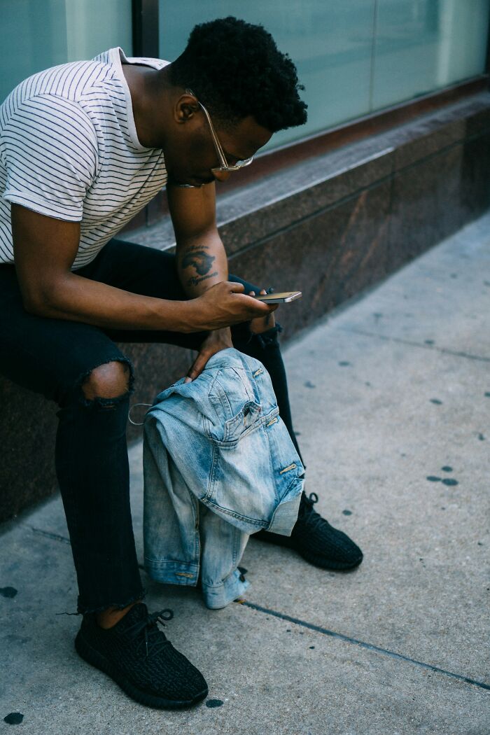 Man sitting on pavement, holding a denim jacket, and looking at his phone after being fired abruptly by his boss.