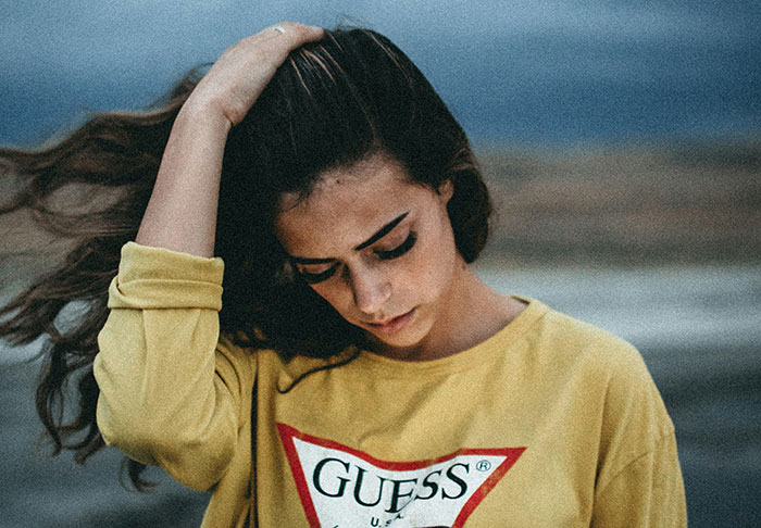 Woman in a yellow Guess sweatshirt, outdoors, with a thoughtful expression, hand in hair.