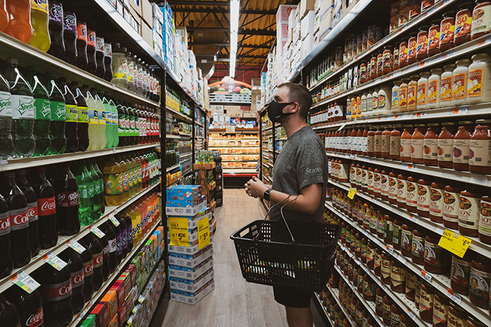 Person wearing a mask holding a basket, standing in a grocery aisle filled with assorted beverage and condiment brands.
