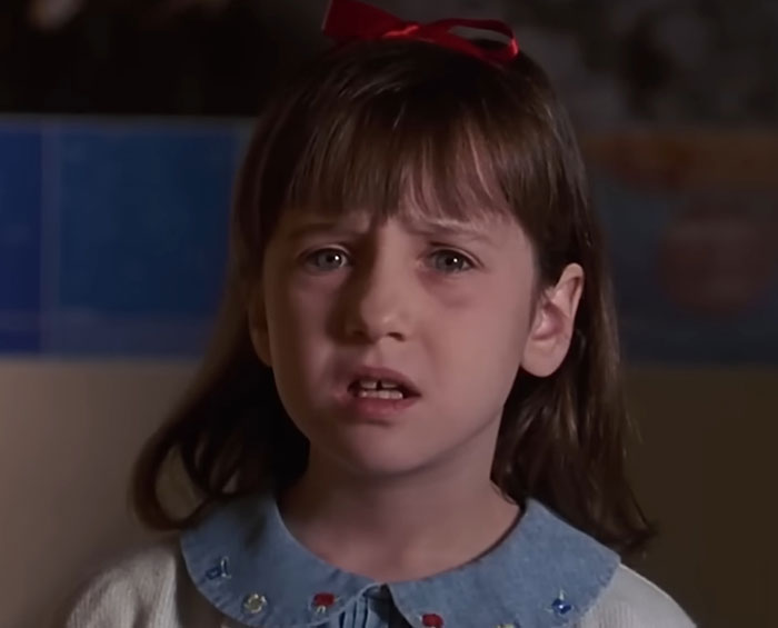 A young girl with a red bow in her hair looks concerned, contrasting her wholesome appearance with a disturbing expression.