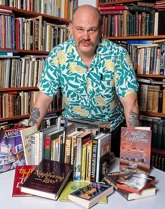 Man in a colorful shirt with tattoos, surrounded by books, standing in a library.