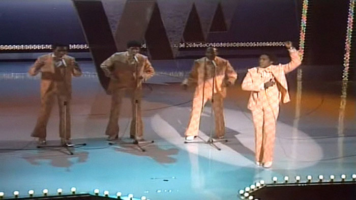 Performers in orange suits singing on stage with vintage microphones, reflecting wholesome vibes with hidden backstories.