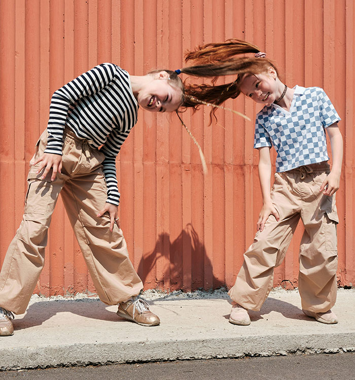Two kids smiling, playfully intertwining long hair, wholesome moment in casual outfits against an orange wall.