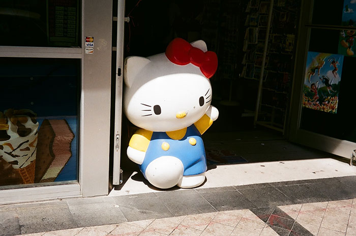 A large Hello Kitty statue, wearing blue overalls, stands by a store entrance.