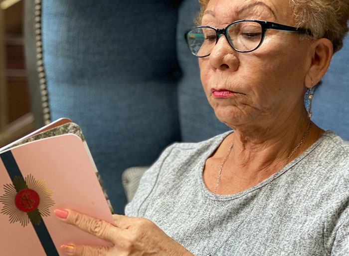 A woman wearing glasses and a gray shirt, deeply engaged in reading a pink notebook, contemplating DNA test results.