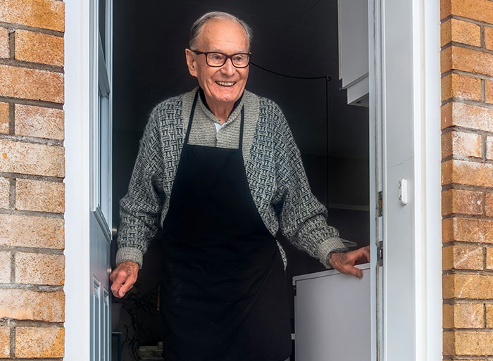 "Elderly man smiling at doorway, wearing glasses and apron, related to DNA test family secrets."