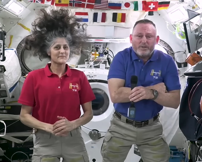 Astronaut with hair floating in zero gravity, wearing a red shirt, inside a space module with flags displayed above.