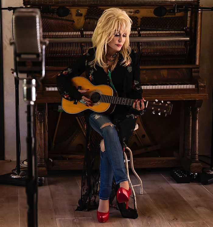 Singer with guitar near a piano, releasing a heartfelt song dedicated to her late husband.