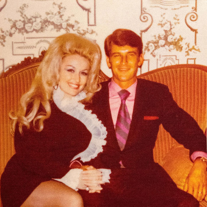Couple sitting on a vintage couch, one wearing a formal dress and the other in a suit, smiling at the camera.