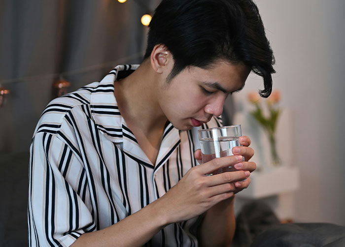 A man in striped pajamas holding a glass of water, sitting indoors by a nightstand with flowers.