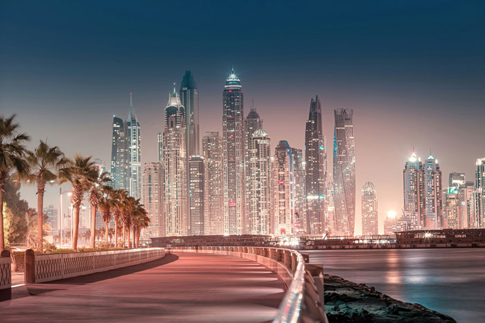Skyline of Dubai at night with lit skyscrapers and a waterfront path.