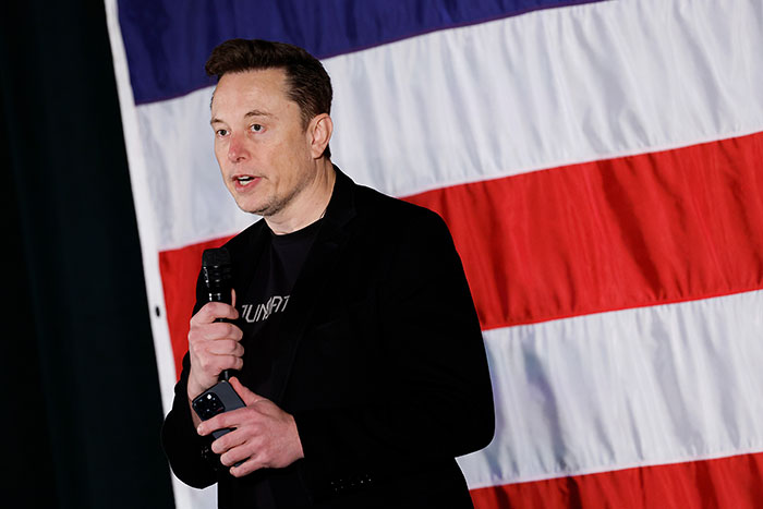 Man speaking with microphone in front of a large American flag, related to SpaceX rescue mission delay. Man speaking with microphone in front of a large American flag, related to SpaceX rescue mission delay.