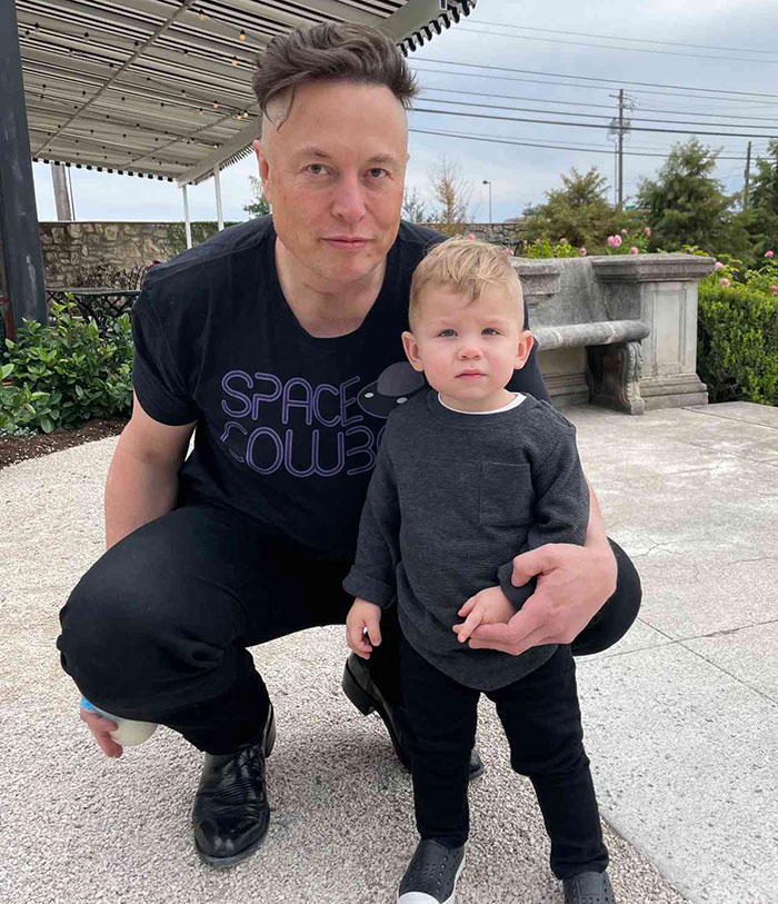 Man in a "Space Cowboy" shirt kneeling beside a toddler outdoors.