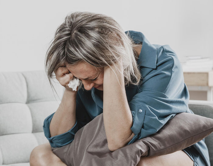 Woman in a blue shirt sitting on a couch, holding a tissue and looking upset after being called out about kids questions. Woman in a blue shirt sitting on a couch, holding a tissue and looking upset after being called out about kids questions.