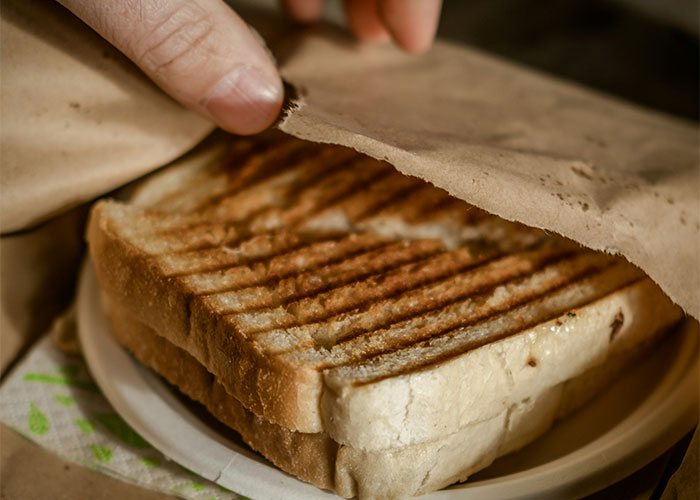 Grilled sandwich in brown paper, partly opened by a hand, possibly in a restaurant setting.