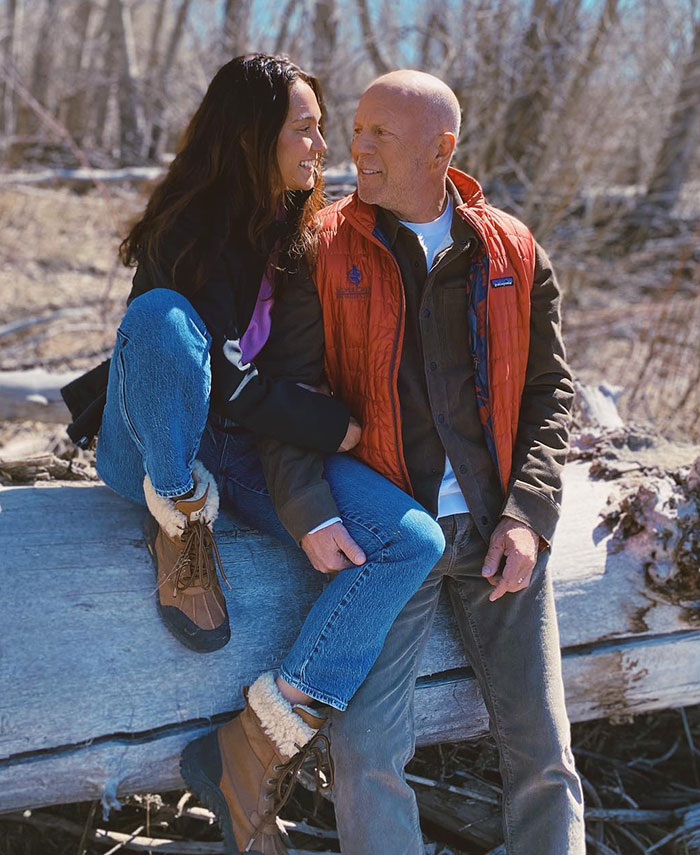 Couple sitting outdoors, man in orange vest, woman in black jacket, nature setting. Keywords: Bruce Willis, dementia. Couple sitting outdoors, man in orange vest, woman in black jacket, nature setting. Keywords: Bruce Willis, dementia.