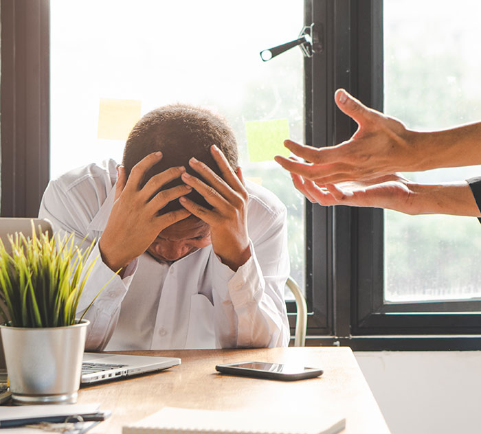 Man in distress at work desk, head in hands, with another's hands gesturing, representing stress from a toxic boss.