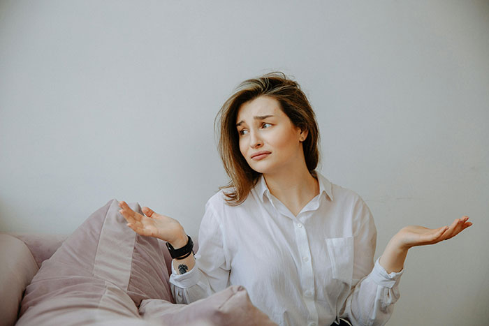 Woman in a white shirt sitting on a sofa, with arms raised and a puzzled expression. Woman in a white shirt sitting on a sofa, with arms raised and a puzzled expression.