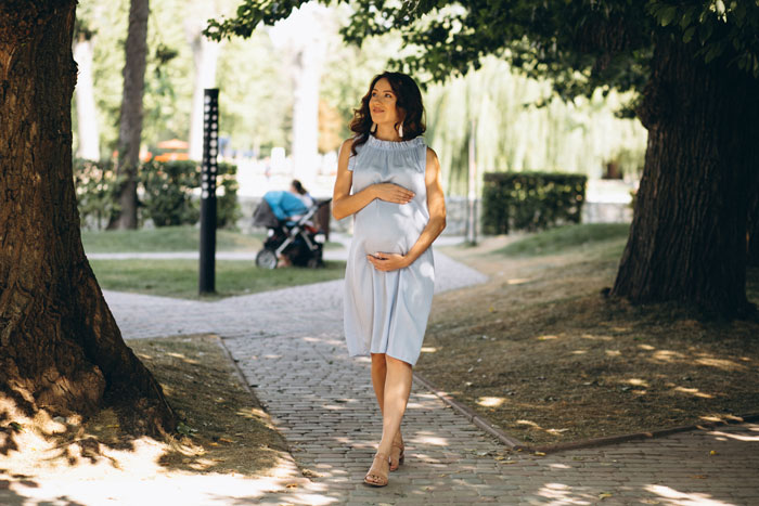 Pregnant woman walking in park, hand on belly, sunny day, trees around.