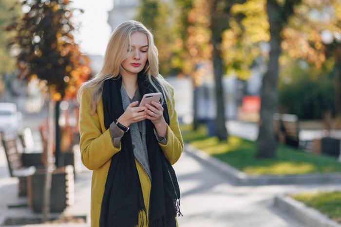 A woman in a yellow coat and black scarf looks at her phone, standing on a tree-lined sidewalk.