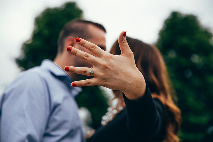 Engaged couple with focus on woman's hand displaying ring, set outdoors with trees in background.