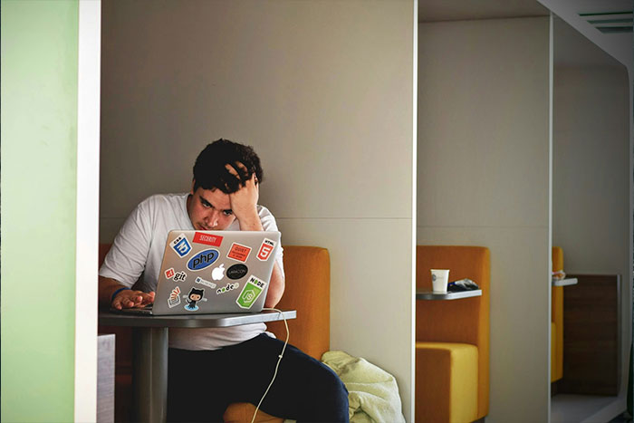 Man looking stressed while using a laptop, covered in tech stickers, in a cafe booth; contemplating ring swap consequences.
