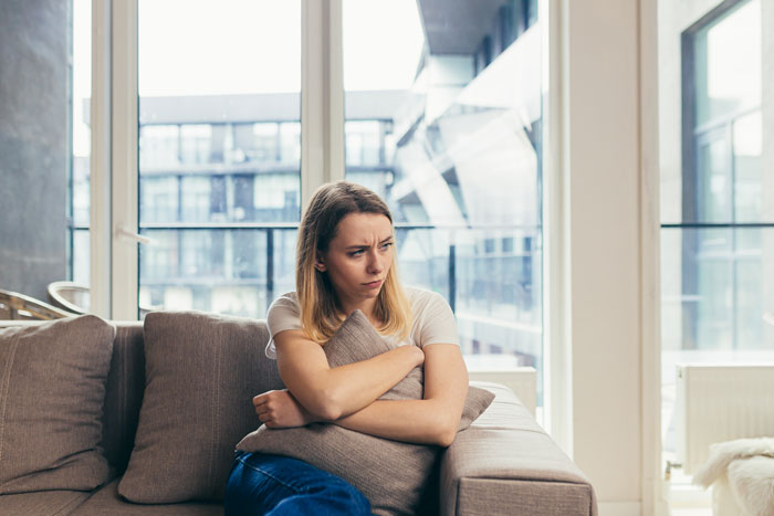 Concerned stepmom sitting on a couch, holding a pillow, pondering her role in her husband&rsquo;s kids' lives.
