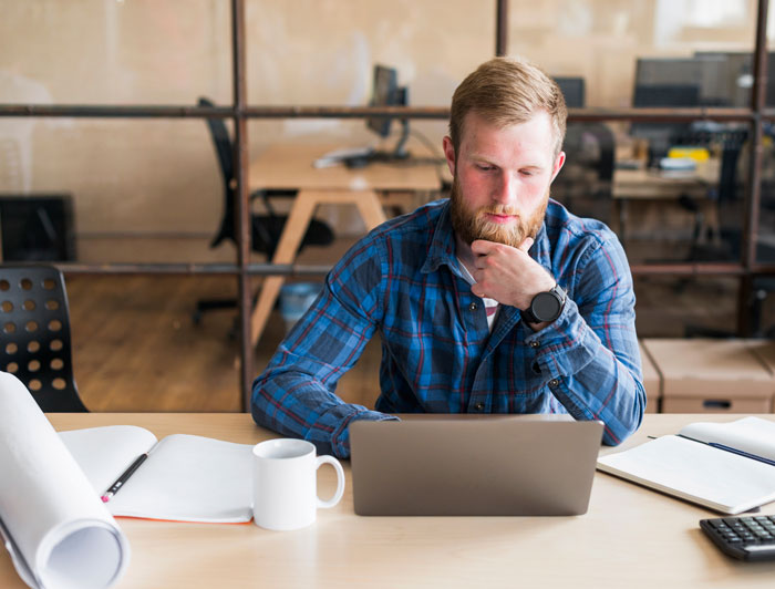 Man in a blue plaid shirt working at a laptop, surrounded by office items, appearing focused.