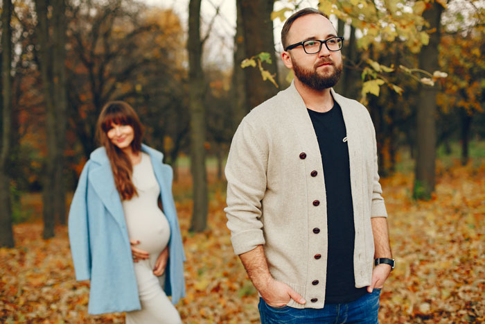 Man in a forest setting, wearing a beige cardigan, with a pregnant woman in the background.