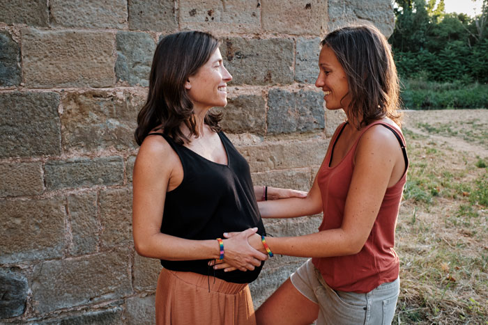 Two women smiling at each other outdoors, one touching the other's belly, symbolizing connection over her partner&rsquo;s baby.