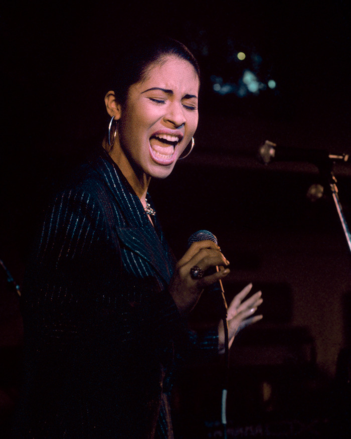 A singer passionately performing on stage, holding a microphone, with eyes closed and wearing a dark outfit.