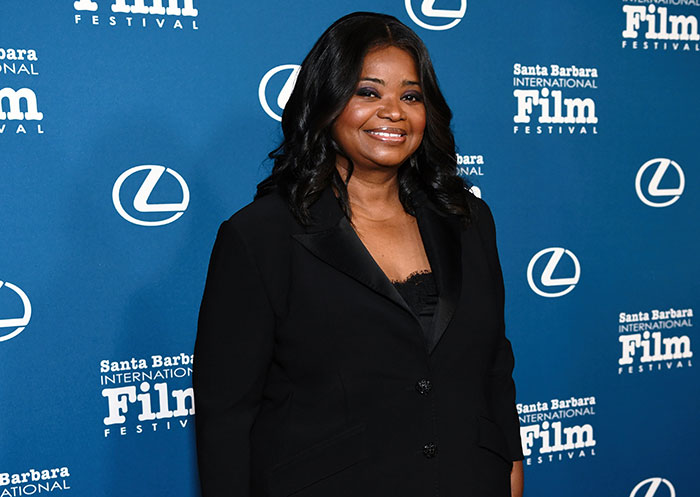 Celebrity woman at film festival in black outfit, smiling against branded blue backdrop.