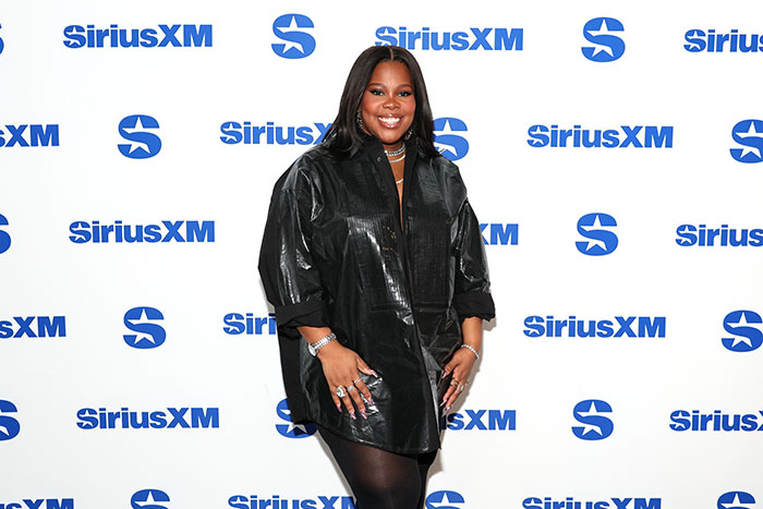 Celebrity at SiriusXM event, wearing a black outfit, smiling confidently despite designer refusals due to weight concerns.