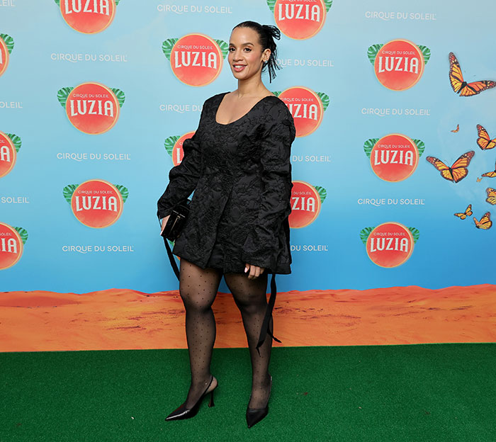 Female celebrity in black outfit at Luzia Cirque du Soleil event, standing on green carpet with colorful backdrop.