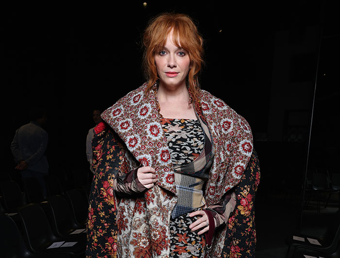 Celebrity woman in a floral outfit, standing confidently at a fashion event amid dim lighting.