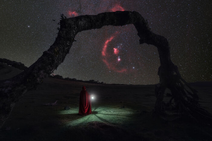 Photographer's stunning landscape of starry sky with a lone figure in red cloak under tree arch.