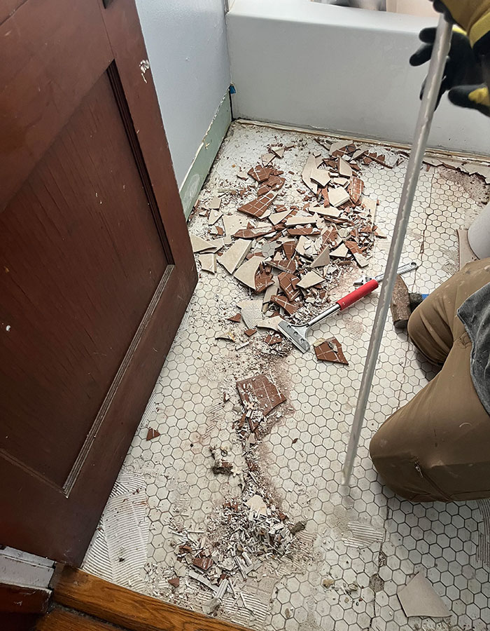 Person uncovering interesting artifacts beneath tile floor in an old house bathroom, using a tool with gloves on.