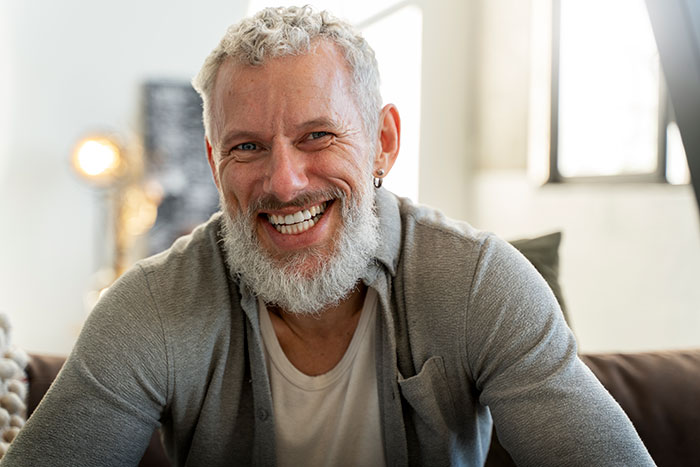Smiling grown man with a beard wearing a gray cardigan, sitting on a couch in a bright room.