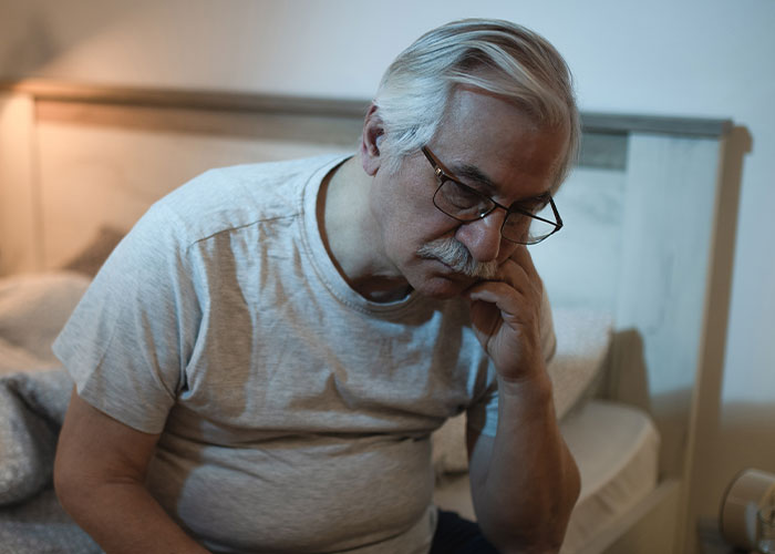 Elderly man sitting on bed, looking pensive and thoughtful, relating to family care.