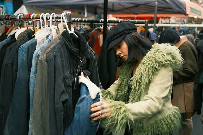 A person browsing clothes at an outdoor market, wearing a unique green furry coat, exemplifying society's acceptance of weird fashion.