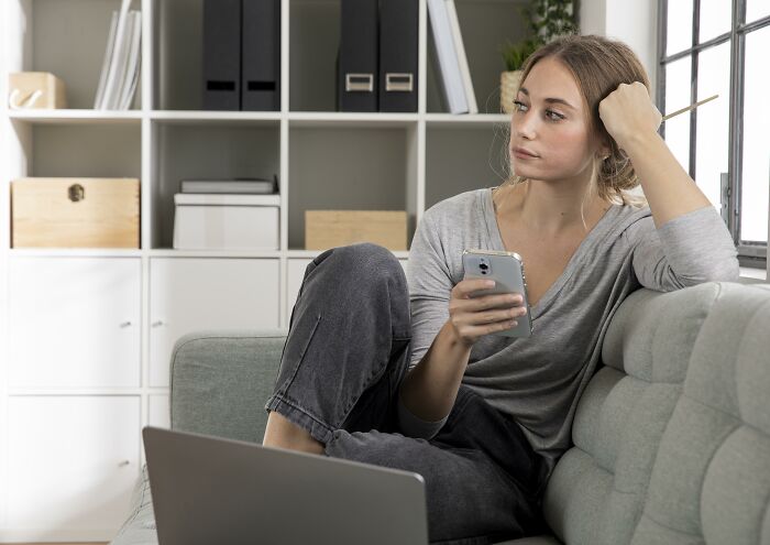 Woman looking concerned, holding a phone while seated on a sofa, shocked by fianc&eacute;'s marital status revelation.
